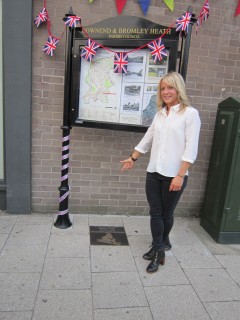 Jenny Jones in front of the Parish Notices board pointing down at her newly unveiled plaque